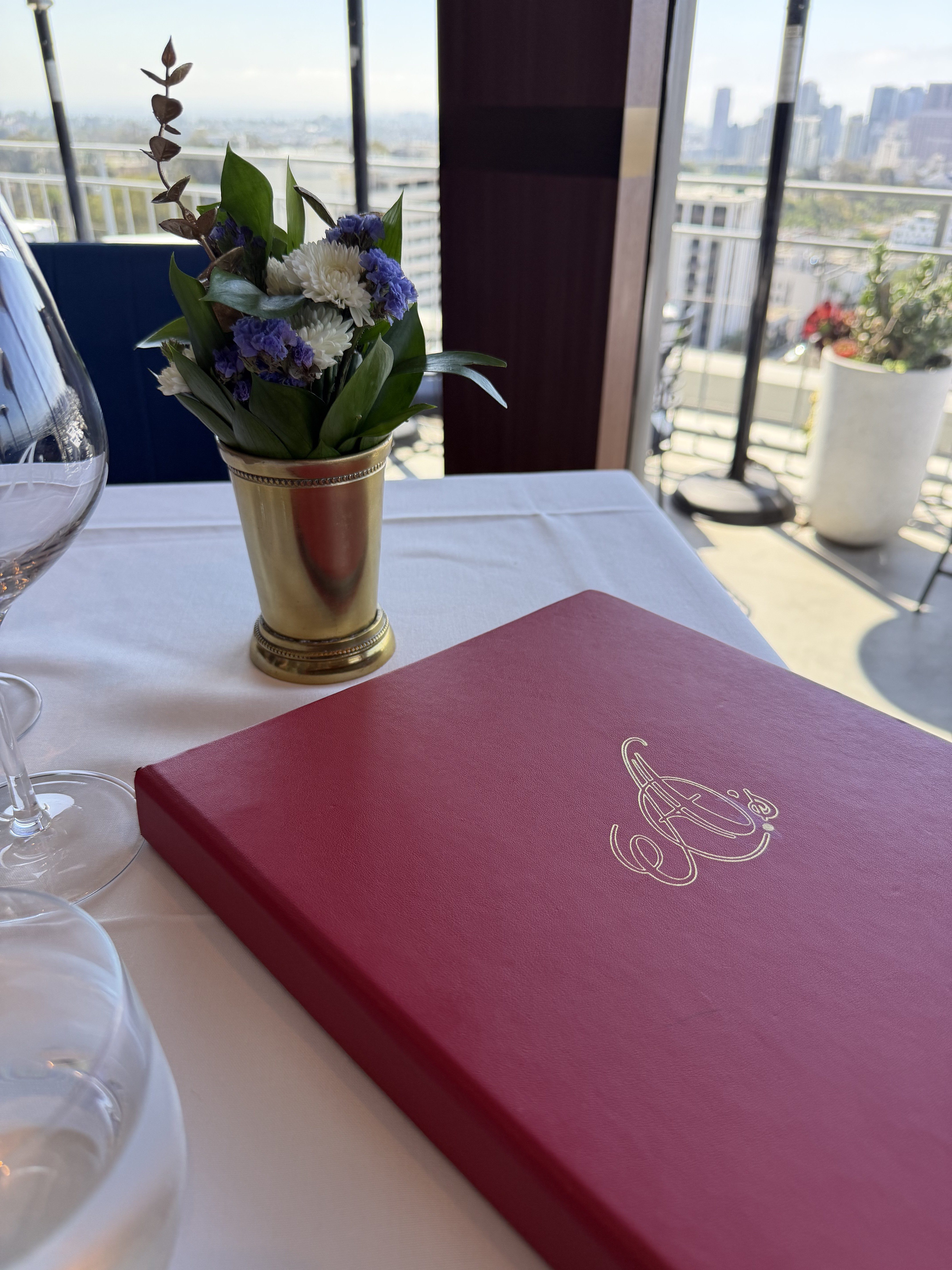 Table set with a red Mister A's menu and fresh flowers against a floor-to-ceiling view of the San Diego skyline